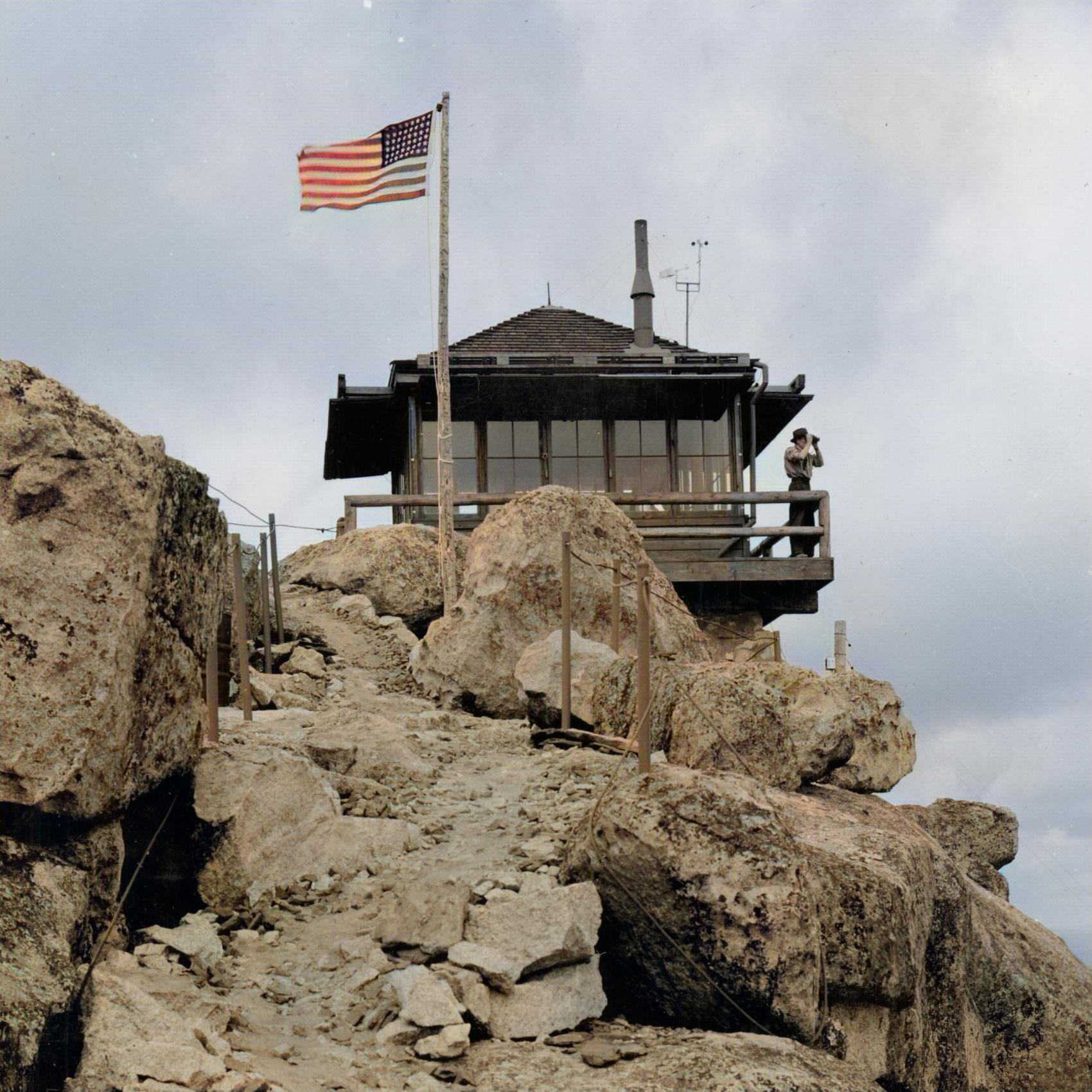 Blank and white photo of a cabin that is standing on a pile of large boulders. A person is looking outside of it to see if there are any fires or anything of note to see. There is an American flag flying by it.